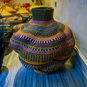 Puede incluir: Una cesta grande tejida con una forma orgánica única. La cesta está hecha de fibras naturales y presenta un patrón geométrico colorido en tonos de verde, morado, amarillo y rosa.