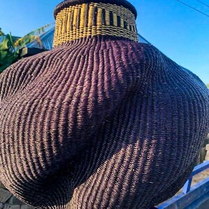 May include: A large, woven basket with a bulbous body and a narrower neck. The basket is primarily dark purple with a textured, ribbed pattern. The neck features a band of yellow and black woven detail, adding a contrasting element to the design. The basket is outdoors against a blue sky.