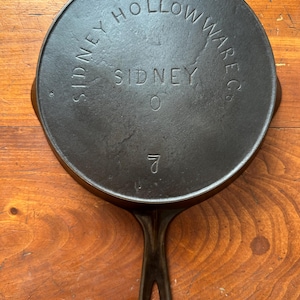 Puede incluir: Una sartén de hierro fundido con acabado negro. La sartén tiene un mango y las palabras "Sidney Hollow Ware Co. Sidney" están grabadas en la parte inferior.