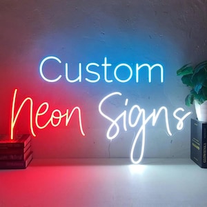 May include: A neon sign with the words "Custom Neon Signs" in white, red, and blue. The sign is illuminated against a light blue and white background. A small plant in a white pot sits on a shelf to the right.