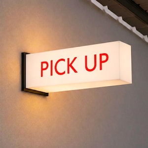 May include: Rectangular illuminated sign with the words "PICK UP" in red letters. The sign is mounted on a dark metal bracket against a light gray wall. The sign is lit from within, creating a soft glow.