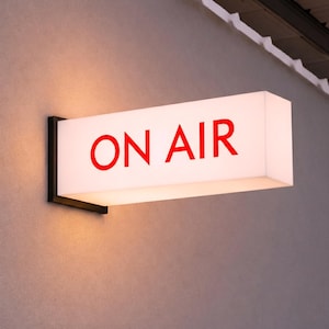 May include: A rectangular, illuminated sign with the words "ON AIR" in red letters. The sign is mounted on a dark metal bracket against a gray wall. The sign is lit, suggesting it is in use.