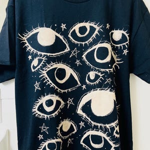 Könnte beinhalten: Schwarzes T-Shirt mit cremefarbenem Augendesign und Sternakzenten. Die Grafik zeigt mehrere stilisierte Augen in verschiedenen Größen und Formen, die ein himmlisches oder mystisches Thema erzeugen. Das Shirt ist ein lässiges Kleidungsstück für den Alltag.