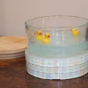 Op de afbeelding: Een heldere glazen kandelaar met een lichtblauwe was binnenkant, met drie gele badeendjes. De basis is versierd met iriserende vierkante tegels. Een houten deksel ligt opzij.