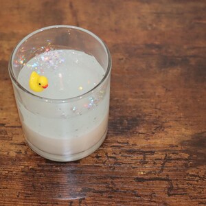 Op de afbeelding: Een glazen pot met een witte kaars erin. De kaars heeft een geel rubberen eendje dat er bovenop drijft en glinsterende glitter door de hele kaars.