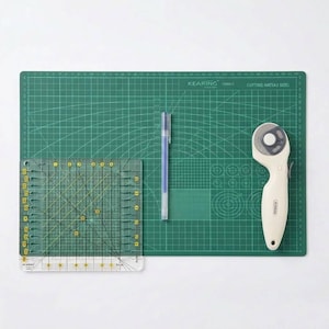 May include: A green A3 size Kearing cutting mat with white grid lines, a clear square quilting ruler with yellow markings, a blue ink pen, and a beige rotary cutter with a circular blade. The mat features various angle and circle guides.