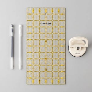 May include: A clear rectangular quilting ruler with yellow grid markings and angle lines, labeled KEARING XPR1206, accompanied by two slim pens with white and black caps, and a white circular suction cup handle tool with the KEARING brand name printed.