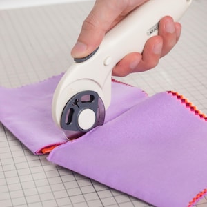 May include: A rotary cutter with a cream-colored handle and a circular metal blade is cutting through a stack of light purple fabric squares. The fabric features a decorative pink and orange zigzag edge pattern along the sides of the stack.