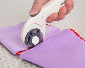 May include: A rotary cutter with a cream-colored handle and a circular metal blade is cutting through a stack of light purple fabric squares. The fabric features a decorative pink and orange zigzag edge pattern along the sides of the stack.