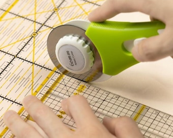May include: A green rotary cutter with a white knob and the text "Gommu Rotary Cutter" is being used to cut fabric on a yellow and white gridded ruler.