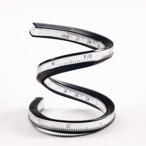 May include: A black flexible ruler with white markings is coiled into a spiral shape. The ruler is marked with numbers from 1 to 10.