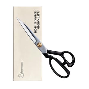 May include: Left-handed fabric scissors with silver-toned metal blades and black plastic handles. The scissors feature a gold-toned pivot screw and are positioned over a rectangular cream-colored box labeled KEARING LEFT HANDED FABRIC SCISSORS.