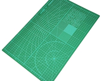 May include: A rectangular green self-healing cutting mat featuring white grid lines, angle markings, and circular guides. The mat is labeled KEARING CM-A3 CUTTING MAT A3 SIZE and includes a ruler along the edges for precise measurements in inches.