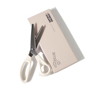 May include: Pinking shears with metallic serrated blades and off-white plastic handles, positioned next to a rectangular beige box. The box features the text "PINKING SHEARS" and a line drawing of the scissors, indicating a 9 inch, 5mm triangle cut.