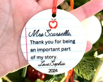 May include: A round white ceramic ornament with a red ribbon, featuring a lined paper design, a red apple icon, and the text: Mrs Scarsella, Thank you for being an important part of my story. Love, Sophia 2024.
