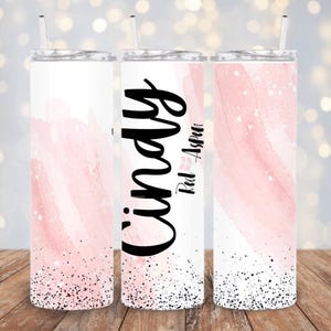 May include: Three stainless steel tumblers with pink watercolor designs and black glitter accents. The tumblers have lids and straws. The middle tumbler has the name "Cindy" and the text "Paul Aspen" written on it.