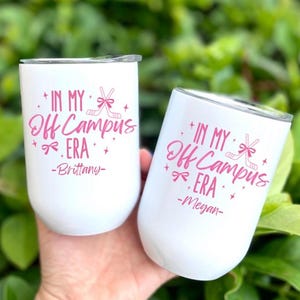 以下が含まれることがあります： 蓋付きの白いステンレス製タンブラー2個。それぞれにピンク色の文字で「IN MY Off Campus ERA」と書かれ、ホッケーのスティックとリボンのグラフィック、そして「Brittany」と「Megan」という名前が記されており、小さな星の模様が添えられています。
