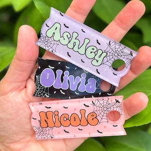 May include: Three personalized acrylic keychains in a hand. The keychains are rectangular with a rounded end and a hole. Each keychain has a name in a different color: Ashley, Olivia, and Nicole. The keychains have a Halloween theme with spiderwebs and bats.