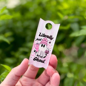 May include: A rectangular acrylic tag with a hole at the top, featuring a light pink and white striped background. The design shows a white ghost wearing a pink beanie, holding a pink tumbler and a pumpkin bucket, with the text Literally just a Ghoul.