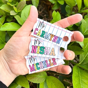 May include: Three colorful teacher name tags with a notebook paper design. The names are "Ms. Carmynn", "Ms. Nisha", and "Ms. Michelle".
