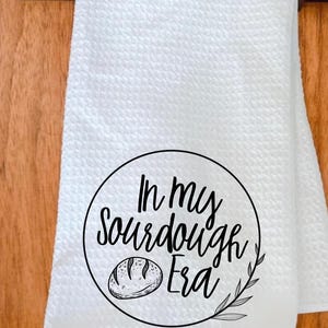 Puede incluir: Toalla de cocina blanca con un patr&oacute;n texturizado. La toalla presenta un dise&ntilde;o circular negro con el texto "In my Sourdough Era" y una ilustraci&oacute;n de pan. Tambi&eacute;n se incluye una ramita de hojas.