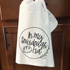 Puede incluir: Pa&ntilde;o de cocina blanco con textura de gofre, colgado de la puerta de un armario. El pa&ntilde;o presenta un dise&ntilde;o circular negro con el texto "In my Sourdough Era" y una ilustraci&oacute;n de pan.