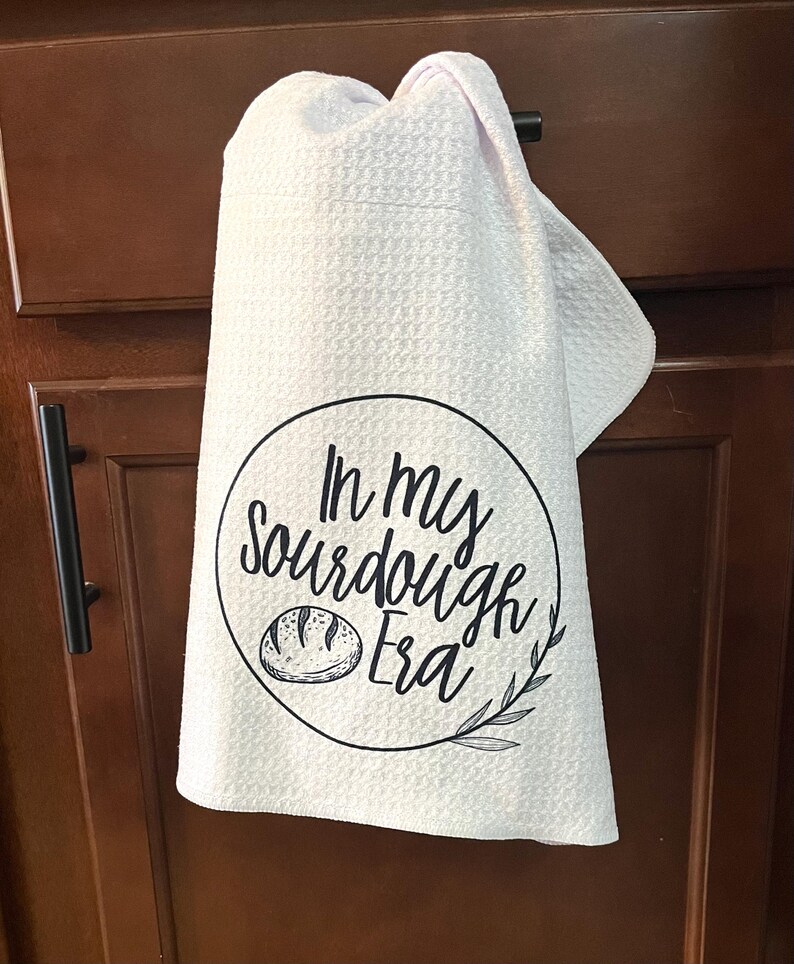 Puede incluir: Pa&ntilde;o de cocina blanco con textura de gofre, colgado de la puerta de un armario. El pa&ntilde;o presenta un dise&ntilde;o circular negro con el texto "In my Sourdough Era" y una ilustraci&oacute;n de pan.