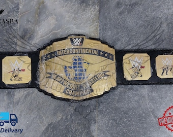 Puede incluir: Un cinturón negro y dorado del Campeonato Intercontinental de Lucha Libre de Peso Pesado. El cinturón presenta placas doradas con el logotipo de la WWE y las palabras "Intercontinental Heavyweight Wrestling Champion". El cinturón está sobre una superficie gris.
