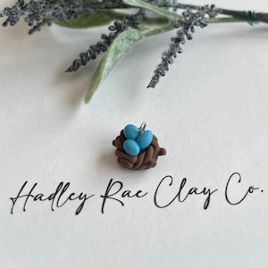 May include: A small brown clay bird's nest with three blue eggs inside. The nest has a silver loop for hanging. The nest is on a white background with the text "Hadley Rae Clay Co."