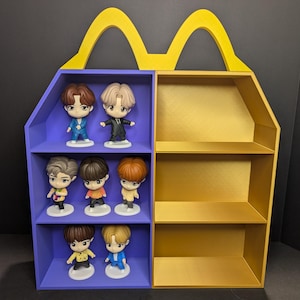 Op de afbeelding: Een dubbelzijdig displayrek in de vorm van een fastfoodcontainer. De linkerkant is paars met drie planken met zes kleine beeldjes. De rechterkant is goudkleurig met drie lege planken. De bovenste handgreep is goudkleurig.