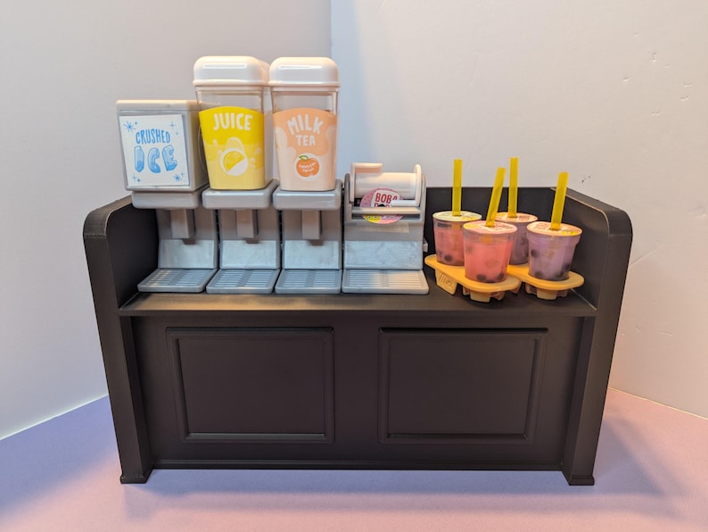 May include: A miniature black beverage stand with dispensers labeled "Crushed Ice", "Juice", and "Milk Tea". Several cups of bubble tea with yellow straws sit on a tray. The stand has a dispenser for boba.