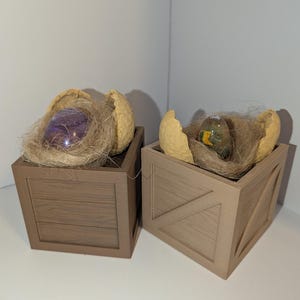 Pode incluir: Duas caixas decorativas de madeira, cada uma com um ninho de barbante e uma casca de ovo rachada. Um ovo é roxo, o outro é verde e amarelo. As caixas têm uma textura de grão de madeira.