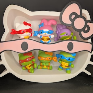 May include: A collection of six figurines, each a hybrid of Hello Kitty and Teenage Mutant Ninja Turtles. The figures are arranged in a white, Hello Kitty-shaped container with a pink bow. The turtles are green, and the other characters are white, pink, and purple.
