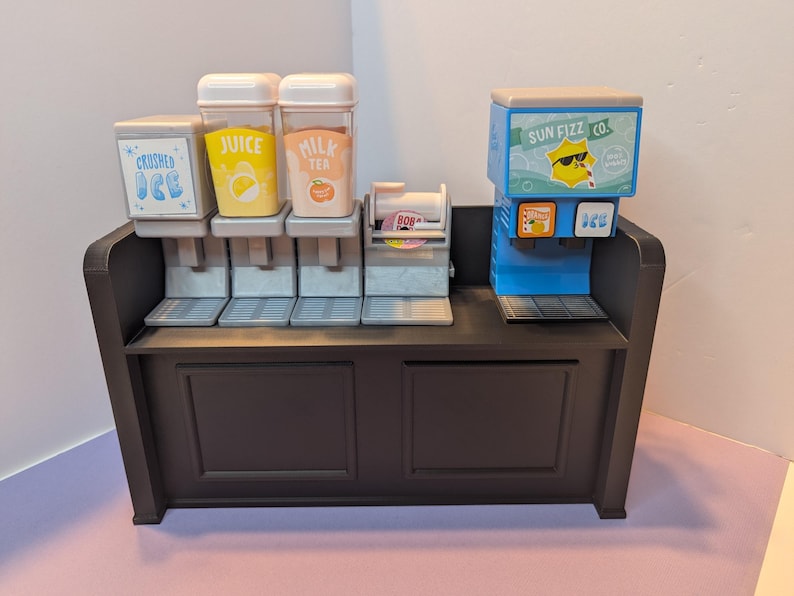 May include: A miniature beverage station with a dark brown cabinet and multiple dispensers. The dispensers include options for crushed ice, juice, milk tea, and a Sun Fizz Co. soda machine. The station is designed with a playful, cartoonish aesthetic.