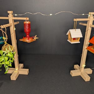 May include: Wooden bird feeder display with various feeders and a hanging plant. The display features a red hummingbird feeder, a small birdhouse, and a teal bird feeder. The display is lit with small fairy lights.