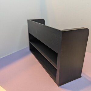 May include: A dark brown, two-shelf display shelf with a rectangular shape. The shelf has a back panel and rounded corners. The unit is placed on a pink and purple surface, with a white wall in the background.