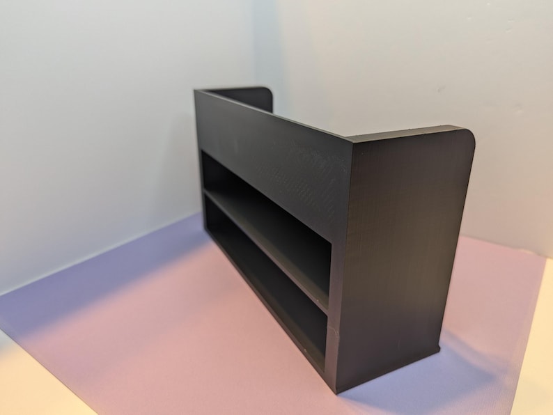 May include: A dark brown, two-shelf display shelf with a rectangular shape. The shelf has a back panel and rounded corners. The unit is placed on a pink and purple surface, with a white wall in the background.