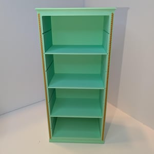 Puede incluir: Una estantería de cuatro baldas con un acabado verde claro y un ribete de cuerda dorada. La estantería tiene un diseño sencillo con líneas rectas y una forma rectangular.