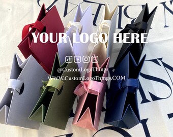 May include: An assortment of colorful paper gift bags in various shades, including red, gray, green, pink, navy, white, and black. Each bag has a handle and is arranged in a group. The text "YOUR LOGO HERE" is displayed on the bags.