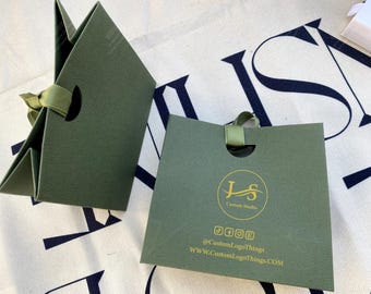 May include: Two olive green gift boxes with a ribbon closure. One box is open, revealing the interior, while the other is closed. The boxes have a circular logo and social media handles printed in gold.