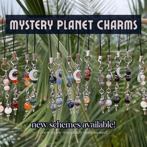 Puede incluir: Una colección de Mystery Planet Charms, cada uno con un diseño único. Los amuletos presentan una variedad de cuentas coloridas, medias lunas y acentos en forma de estrella. El texto "Mystery Planet Charms" se muestra encima de los amuletos, con "new schemes available!" debajo.