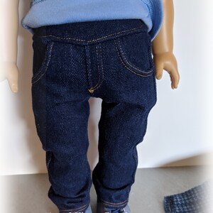 May include: A pair of dark blue denim jeans for a doll, with a light blue shirt and grey shoes. The jeans have a classic five-pocket style and a straight leg.