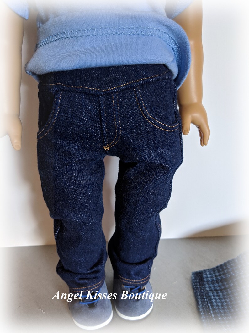 May include: A pair of dark blue denim jeans for a doll, with a light blue shirt and grey shoes. The jeans have a classic five-pocket style and a straight leg.