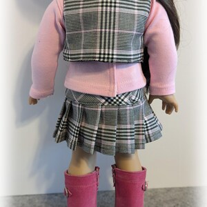 Op de afbeelding: Een roze en zwart geruite rok en vest set voor een pop. De rok is geplooid en het vest heeft een kraag. De pop draagt een roze longsleeve shirt en roze laarzen.