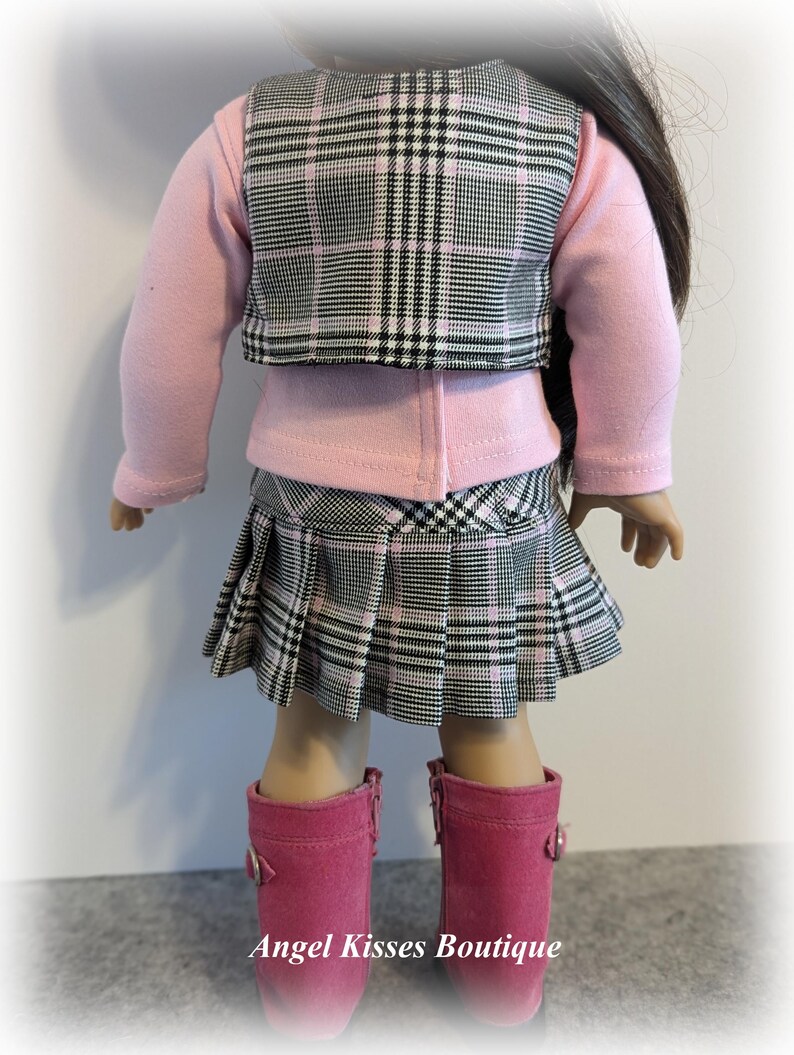 Op de afbeelding: Een roze en zwart geruite rok en vest set voor een pop. De rok is geplooid en het vest heeft een kraag. De pop draagt een roze longsleeve shirt en roze laarzen.