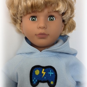 May include: A doll with blonde hair and green eyes wearing a light blue hoodie. The hoodie features a patch of a blue video game controller with a yellow lightning bolt and a plus sign. The text "Angel Kisses Boutique" is at the bottom.