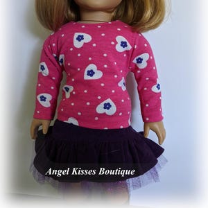 May include: A pink long-sleeved shirt with white hearts and blue flowers, paired with a dark purple skirt and a tulle overlay. The shirt has a polka dot pattern. The skirt has a ruffled hem. The text "Angel Kisses Boutique" is visible at the bottom of the image.