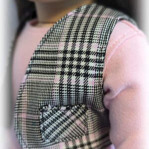 Op de afbeelding: Een zwart-wit geruit vest met roze accenten. Het vest heeft een klein zakje aan de voorkant. Het vest wordt gedragen over een roze shirt.