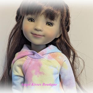 May include: A doll with long brown hair wearing a pastel tie-dye hoodie. The doll has brown eyes and is smiling.
