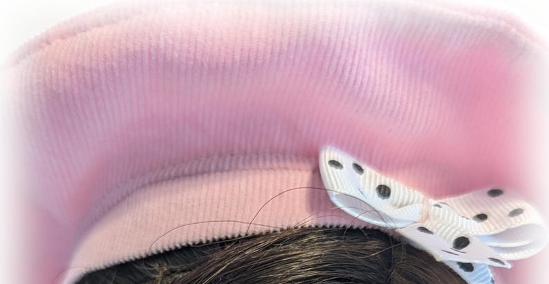 Op de afbeelding: Een roze corduroy baret met een witte strik versierd met zwarte stippen.